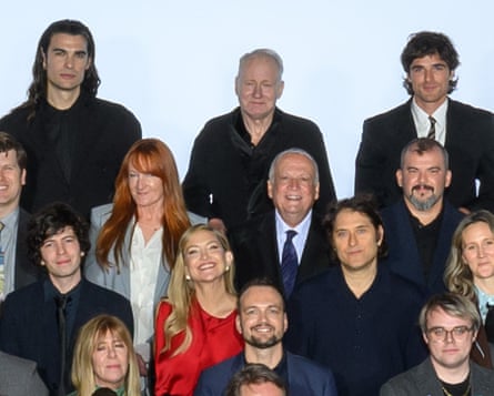 At the back of a group shot, a tall man with long, dark hair is shown standing next to an older gentleman who has a dark haired man next to him.