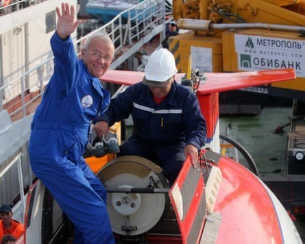 An elderly white-haired man in a blue boiler suit climbs into a submarine, out of the water, while a man in a hard hat helps him