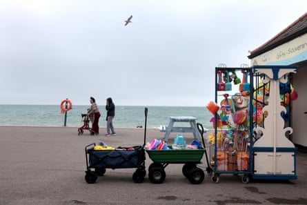 Bognor Regis: a seafront kiosk sells buckets, spades and beach toys on a cloudy day; two people, one with a walker, are on the promenade and the sea looks grey but calm in the background; a seagull flies overhead.