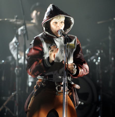 Robyn in a hooded jacket sings into a microphone on stage.