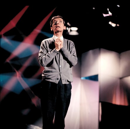 Kenneth Williams in a grey cardigan, hand clasped together, under the lights of a TV stage set