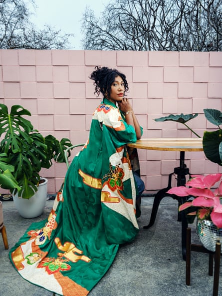 Moni Sebestyen. Portrait of Simone Ledward Boseman in a long colourful coat-dress, pictured outside surrounded by sculptural plants