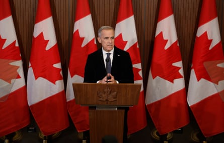 Canada’s prime minister, Mark Carney, delivers a statement from a wooden podium with a line of furled Canadian flags behind him.