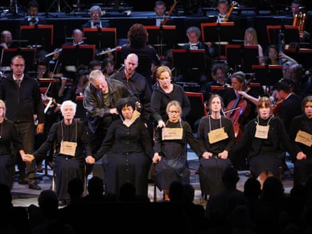 female cast members of the dialogues of the carmelites, dressed in long black nun-like dresses, seated on the royal albert hall stage as other cast members tie signs around their necks. the orchestra is behind them, playing
