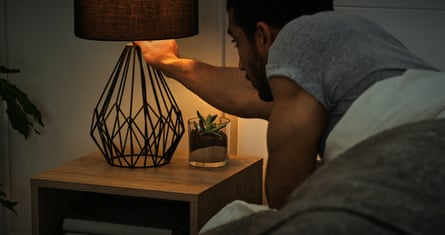 A white man leans over out of bed to turn off a rather chic bedside lamp.