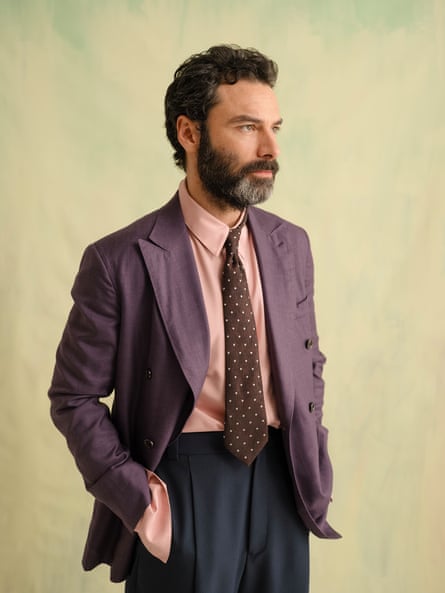 Aidan Turner wearing a purple blazer, salmon shirt, brown tie with a white pattern and baggy navy trousers facing slightly to his left, standing with his hands in his pockets against a mottled pale yellowy-green background