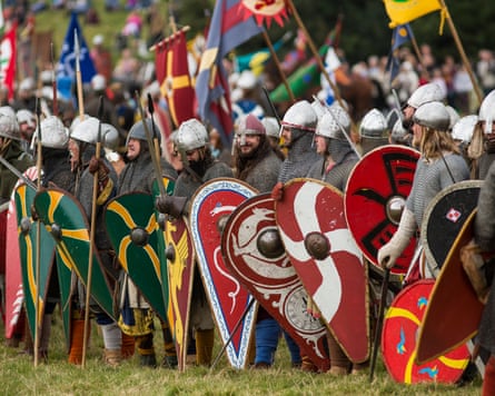 A group of people dressed in medieval combat wear holding vibrant red and green shields and raising flags