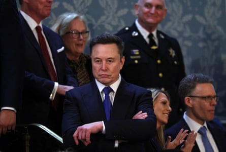 A man in a tight suit stands with his arms crossed in an imperious manner, as if he were channeling Nero at the Colosseum
