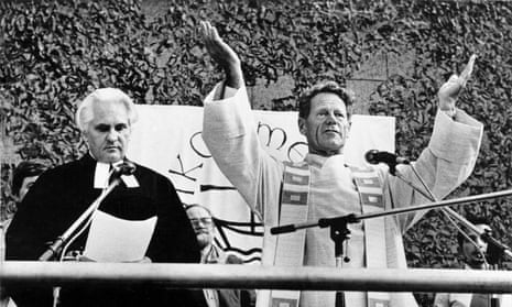 Hans Kung in priest's robes, both arms upraised, with a banner of the ecumenical movement visible behind and another priest standing next to him