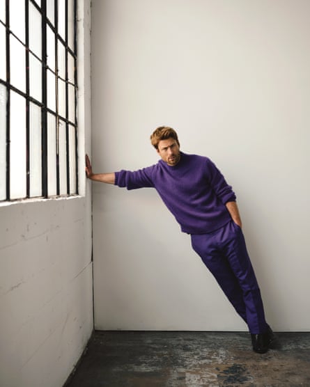 Portrait of Glen Powell wearing deep purple jumper and trousers and black shoes standing away from a white wall with black window frames leaning towards the wall with his right arm full stretched out to balance his weight