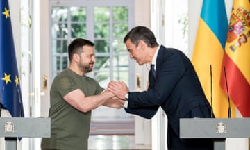 Two men lean towards each other from behind their lecterns and grasp hands. Behind them are the flags for Ukraine and Spain