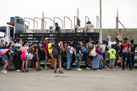 People stand in line by a tall concrete wall, some carrying bags and rucksacks.