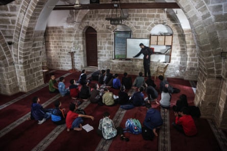 Children sit on mats on the floor of a brick room watching a man standing at a whiteboard
