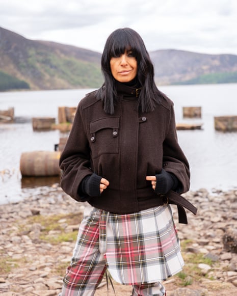 Claudia Winkleman stands in a dark jacket and tartan skirt by a lake with mountains behind