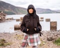 Claudia Winkleman stands in a dark jacket and tartan skirt by a lake with mountains behind