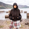 Claudia Winkleman stands in a dark jacket and tartan skirt by a lake with mountains behind