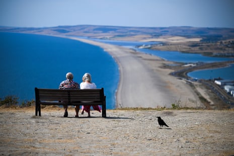 A man and a woman sit on a park bench overlooking a narrow strip of sand between the ocean and a small inlet. A small bird sits in the foreground.