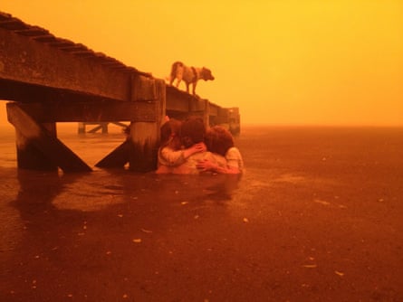 A woman standing in water up to her shoulders holds her grandchildren as a wildfire rages and turns the sky orange