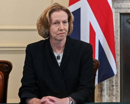 Meg O’Neill sits at a meeting table at Downing Street in front of a union jack flag. She has collar-length blond hair, wears a black jacket, and looks stern.