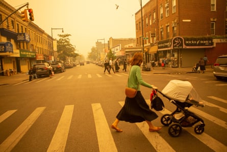 A lady pushes a buggy across a zebra crossing of a New York street with an orange haze in the sky.