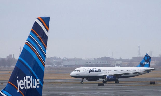 An A320 Jet Blue plane with blue and light blue tail livery taxis at JFK airport in New York. A blue, orange and white striped tail from a Jet Blue plane is seen in the foreground.