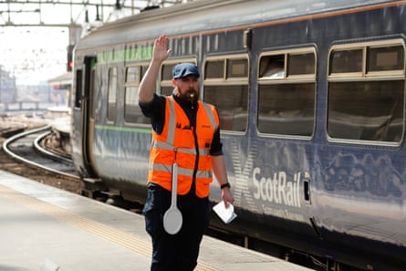 A train guard in an orange hi-vis vest blows his whistle for departure as a Scotrail train departs from Glasgow Central Station.