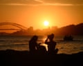 Two silhouetted people watch the sunset from rocks with Sydney Harbour Bridge in the background