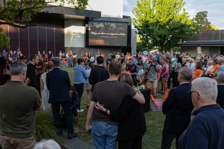 A crowd gathers at a vigil facing a large screen, with a couple embracing in the foreground