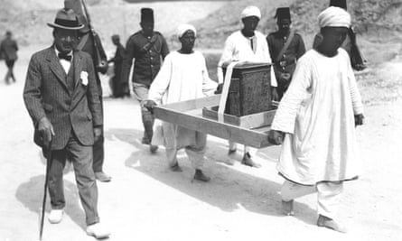 Man in a suit and hat, with a walking stick, accompanies three others carrying a piece of furniture strapped onto a large tray. Other men in uniform walk behind them