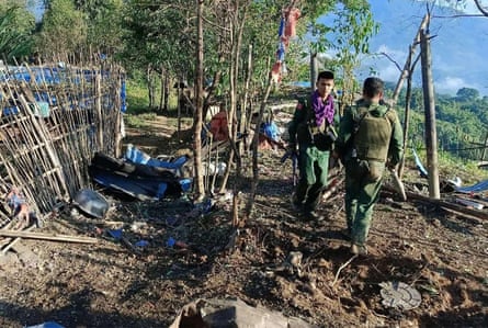 Two men in military fatigues, one with an AK-47, by the remains of huts and a fence