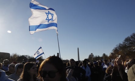 Blue and white Israeli flag with sun shining through it above a crowd.