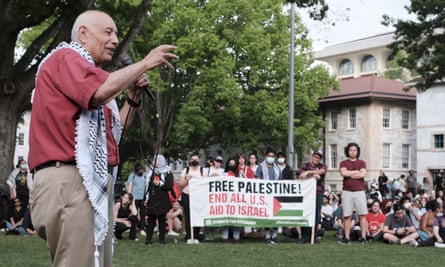 An older Palestinian man wearing a red shirt and khakis and draped in a keffiyeh, holds a microphone and speaks to people gatherd on a lawn.