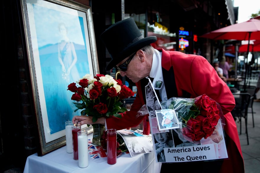 A man in a red coat and top hat lights a candle on a small table that holds several other candles, flowers and a photograph of Queen Elizabeth.