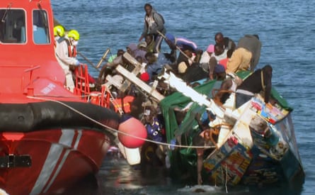 A red coastguard vessel floats alongside a wooden boat which is tipped sideways with poeple scrambling to find safe positions.