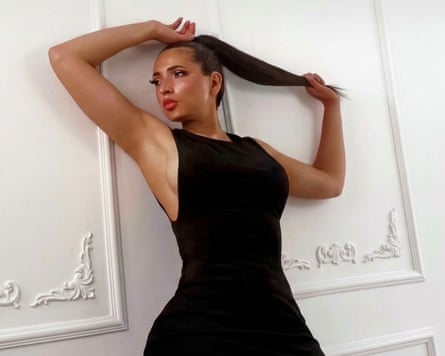Natalia Villalba poses for a photo with one hand raised above her head and the other holding the end of her long, dark hair which is in a slicked-back ponytail. She is wearing a sleeveless, tight-fitting black dress and leans against a white wall or door with carved wooden panelling.