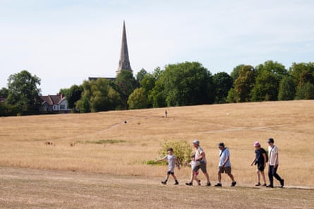 A group of people walk through a parched area of grassland with with trees and a church in the background