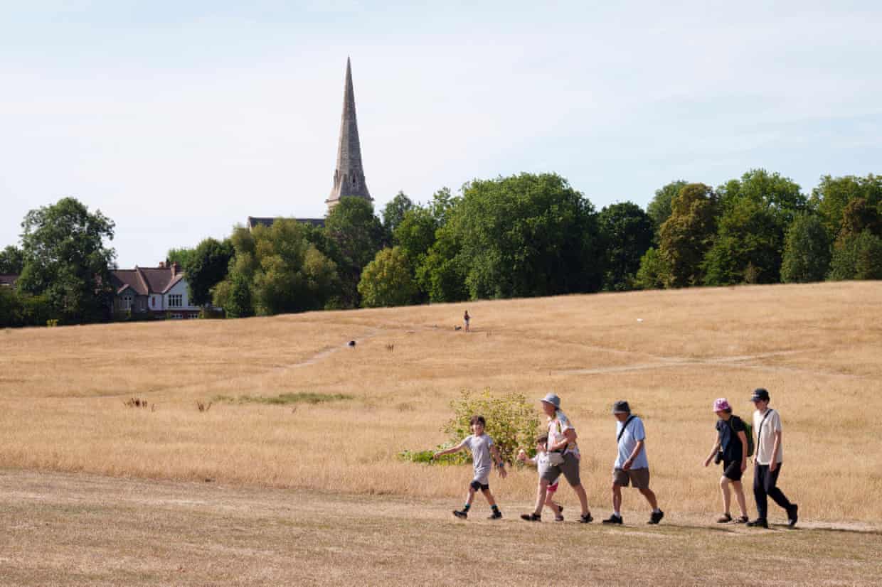 A group of people walk through a parched area of grassland with with trees and a church in the background
