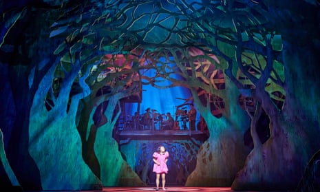 still image from stage performance of My Neighbour Totoro: a girl in a pink dress is seen in the centre of a mythical forest with trees curving around her, lit in green, blue and purple