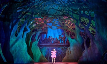 still image from stage performance of My Neighbour Totoro: a small girl in a pink dress is seen in the centre of a mythical forest with trees curving around her, lit in green, blue and purple
