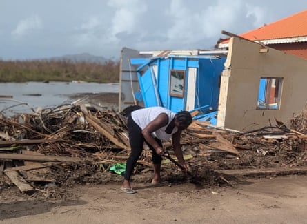 A woman bends down with a shovel to clear dirt and debris next to a damaged house
