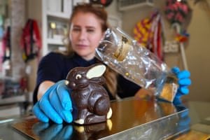 A woman wearing blue protective gloves holds a chocolate rabbit in one hand and a plastic bag in the other, which she is about to place the rabbit in