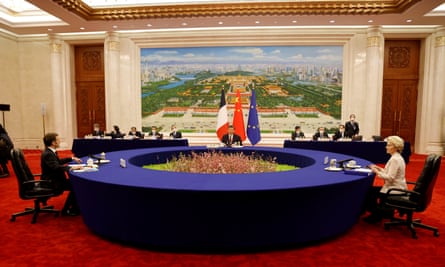 Xi Jinping with Emmanuel Macron and Ursula von der Leyen: they are sitting around a large dark blue circular table at 9 o’clock, 12 and 3 o’clock respectively. The table is set on a bright red, deep carpet and it has a sunken green centrepiece with plants in the middle; Xi sits in front of flags of their three countries and a backdrop of a stylised cityscape; there are panels of other people seen seated at long tables in the background. Given the size of the table and the distance between them, both Macron and von der Leyen look tiny, especially Macron, and the overall effect is quite comical