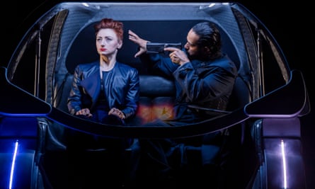 Jodie McNee sitting in a futuristic-looking vehicle next to Ricardo Castro, who has a gun pointed at her