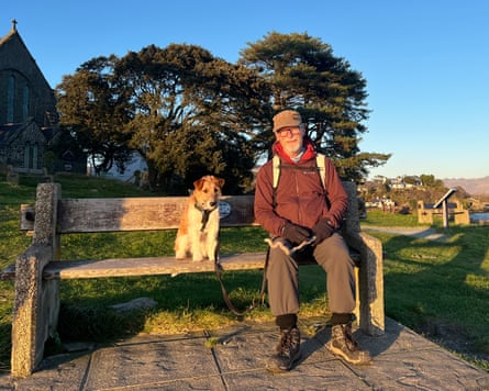 Man in hiking clothes with dog on a public bench. River in background and steep hills