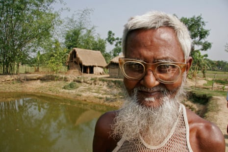 A Bangladeshi man wearing a pair of glasses smiles at the camera. Behind him is a fishpond and fields.