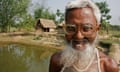 A Bangladeshi man wearing a pair of glasses smiles at the camera. Behind him is a fishpond and fields.