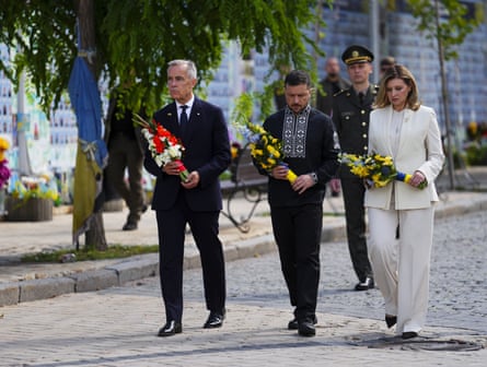 Mark Carney, Volodymyr Zelenskyy and Olena Zelenska walk in a row and carry bouquets of flowers in their national colours
