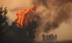Partly obscured by dark gray smoke are silhouettes of about ten firefighters standing on a two-lane road pointing a hose at an orange conflagration in the trees alongside it.