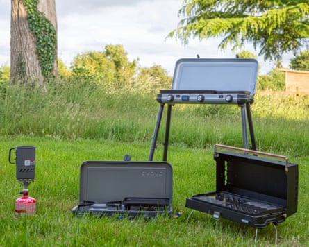 A group shot of four camping stoves in a green field with the sky and trees in the background