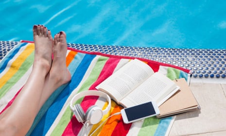 Crossed legs on a colorful towel with books and headphones connected to a smartphone at the edge of the swimming pool